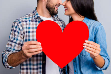 Cropped close up portrait of red paper heart with profile half face smiling couple, nose to nose, face to face, holding love symbol, standing over grey background, 14 februaryの写真素材