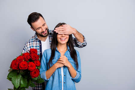 Handsome man with bristle in  shirt making  surprise for his beautiful, charming, joyful lover in casual outfit, closing her eyes with hands over grey background, women's dayの写真素材