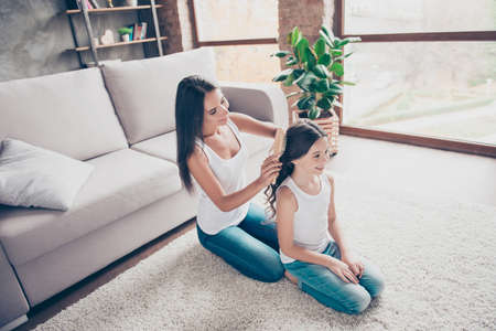 Happy attractive young mother in white tshirt is brushing her little schoolgirl daughter's black long dark hair and making a ponytail, they are sitting on the floorの写真素材