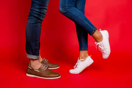 Closeup photo of woman and man legs in jeans, pants and shoes, girl with raised leg, stylish couple kissing during date, isolated over red background, he vs sheの写真素材
