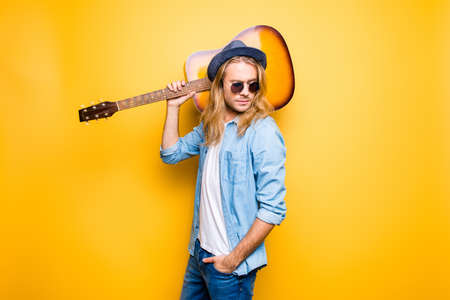Portrait of attractive, bearded guy holding guitar on shoulder, hand in pocket, standing over yellow backgroundの写真素材