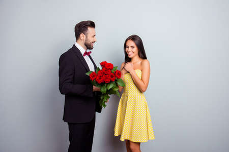 Bearded gentleman in tux with bowtie presenting bouquet of red roses to his charming cute sexy girlfriend in yellow dress on 14 february, wife and huwband on grey backgroundの写真素材