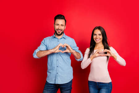 Love story of cheerful, attractive, lovely, cute, sweet couple in casual outfit making hearts with fingers over red backgroundの写真素材