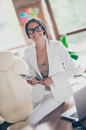 Nice, independent, cute woman in white suit, formal wear, spectacles sitting on desktop in workplace, holding tablet in hands, looking at cameraの写真素材