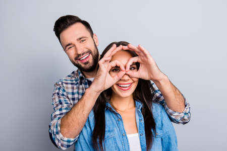 Handsome, bearded man making eyewear with fingers to his wonderful, smiling girlfriend, cheerful couple looking at camera, standing over grey backgroundの写真素材