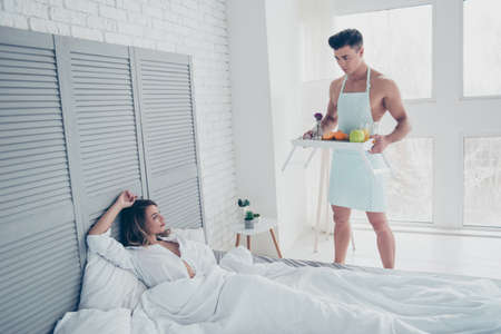 Handsome, attractive, muscular, naked man in apron prepare breakfast in bed for his lover, holding tray with food, pretty woman laying and looking at him, concept of dream on 14 february and 8 marchの写真素材