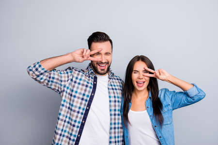 Cheerful, laughing, comic, positive, family couple in shirts embracing and gesturing v-sign near eyes, having one eye closed, standing over grey backgroundの写真素材
