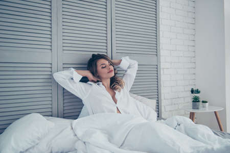 Rest, sleeping, comfort and people concept - young woman stretching in bed in light room at home bedroom, hands behind the head, dreaming about breakfast in bedの写真素材