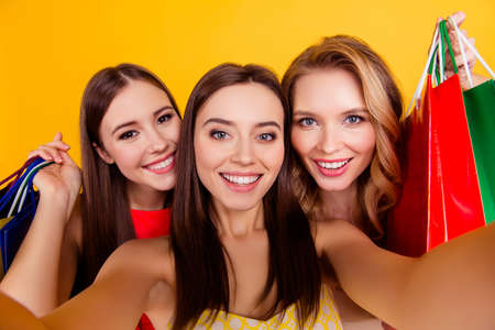 Pretty, charming, stylish girl making selfie photo using front camera with her friends after shopping, holding colorful bags, spending a lot of money, crazy sales, shopaholic, yellow backgroundの写真素材