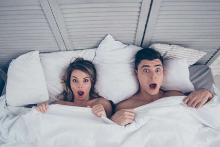 What we did last night? Top view of funny man and woman lying in bed covered by sheets with wide opened eyes and mouth looking at camera, having shock, confuseの写真素材