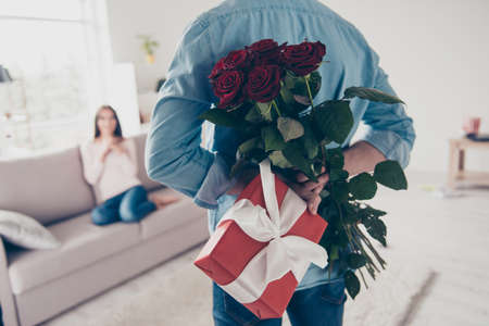 Unexpected moment in routine everyday life! Cropped photo of man's hands hiding holding chic bouquet of red roses and gift with white ribbon behind back, happy woman is on blurred backgroundの写真素材