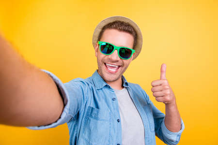 Self portrait of cheerful, bearded blogger in summer glasses shooting selfie, showing v-sign to the camera, making video for his blog over yellow backgroundの写真素材