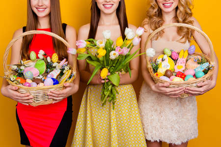 Cropped half face portrait of three attractive, pretty girls holding bouquet of tulips and wicker baskets with colorful traditional easter sweets, gingerbread, eggs, standing over yellow backgroundの写真素材