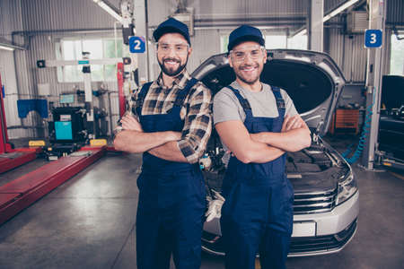 Welcome to our car service. Two experts  with arms crossed, smiling at camera, in special safety outfit and glasses, headwear, standing over background of entrance of automobile in work stationの写真素材