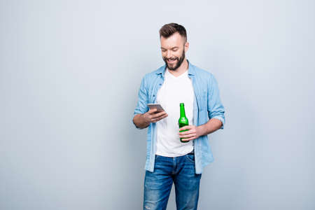 Happy joyful cheerful bachelor is drinking beer and using smartphone for typing sms, isolated on grey backgroundの写真素材