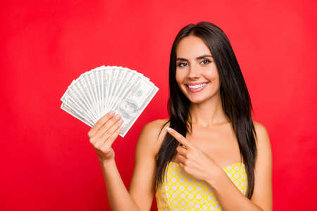 Close up portrait of excited beautiful gorgeous attractive pretty cheerful confident dark-haired woman demonstrating fan made of money and pointing with forefinger isolated on vivid red backgroundの写真素材