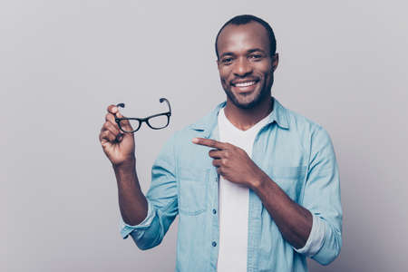 Do you want to try on? Portrait of confident happy smiling joyous cheerful excited afro man wearing casual clothing pointing on glasses in his hand, isolated on grey backgroundの写真素材