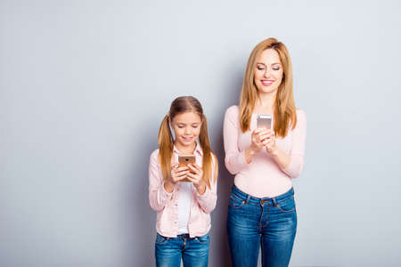 Pretty, charming, nice, mother and daughter over gray background holding digital smart phones in hands, using wifi internet, checking email, chatting, social networkings, appsの写真素材