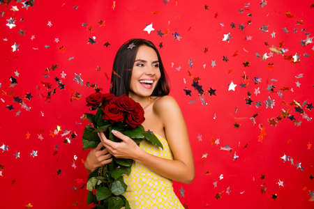 I'm the happiest woman in the world! Portrait of cheerful delightful attractive elegant tender gentle cute lovely luxurious woman with naked shoulders holding enjoying roses isolated on red backgroundの写真素材