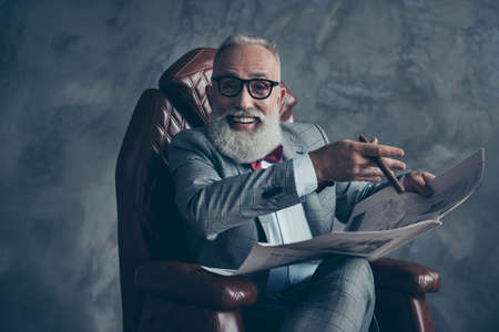 Attractive, rich, old, laughing boss with beaming smile in glasses, tux with red bow, have, hold newspaper, smoke cigarette, sitting in leather armchair in workplace over gray background, relax, funの写真素材