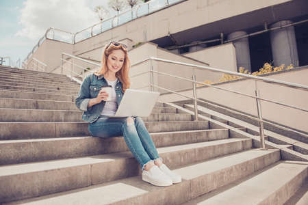 Concept of having free wireless connection. Beautiful cheerful excited woman sitting on stairs in city center clothed in casual fashionable jeans, jacket outfit is sending emails and enjoying teaの写真素材