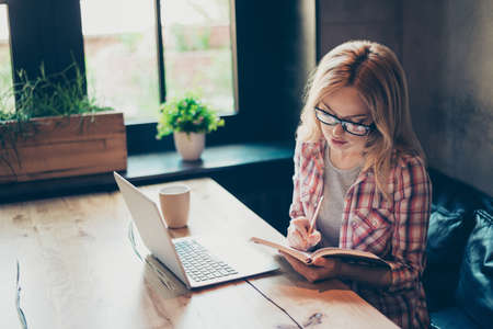 Cute intelligent clever concentrated girl is studying hard in cafe, she is using a laptop with free access to the internet and writing down necessary information to her notebook, sitting at the tableの写真素材
