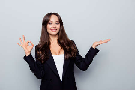 Portrait of brunette, charming, pretty, confident woman in black jacket demonstrating new product with palm on copy space, gesturing ok sign, looking at camera, standing over grey backgroundの写真素材