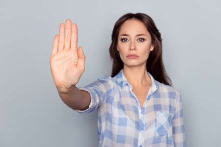 Prohibition symbol. Closeup portrait of young, serious, pretty, charming girl in checkered shirt making stop gesture with palm of her hand on grey backgroundの写真素材