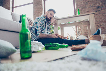 Low angle photo of tired exhausted drunk bearded guy wearing checkered casual shirt and jeans, he is sitting on the floor, sleeping and holding a piece of pizzaの写真素材