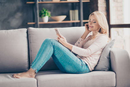 Full length portrait of charming pretty woman in jeans and pullover siting with legs on couch in livingroom, having earphones, using, holding smart phone, listening music with close eyesの写真素材