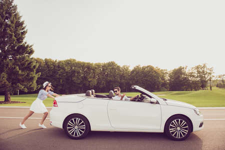 Full length side profile shot of gorgeous brunette lady in cap and brunet guy driver, both well dressed, in black trendy eyewear, resolving the car problem, park with green trees around, sunshineの写真素材