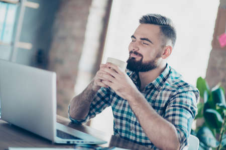 It will be easy for me! Smiling cunning happy cheerful manager wearing checkered shirt is enjoying a cup of fresh hot coffee and having a pause, sitting at the table near laptopの写真素材
