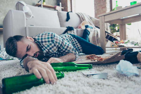 Sick drunk dreamy bearded guy clothed in checkered shirt and denim jeans is sleeping on the floor with an empty bottle in handの写真素材