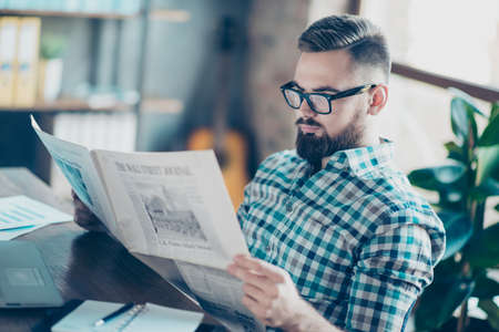 Careless concentrated clever bearded guy clothed in casual checkered shirt is reading a newspaper at work, he has a break, he is sitting at the tableの写真素材