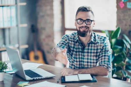 Concept of successful job interview. Portrait of happy cheerful smiling glad worker giving you a hand for handshaking, he is sitting at his comfortable office in front of laptopの写真素材