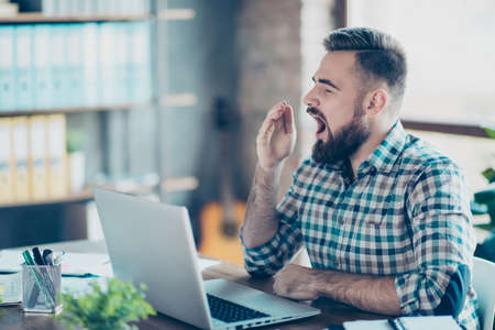 Concept of working day and night without having a recreation. Half-turned photo of exhausted powerless yawning tired young wearing casual checkered shirt office worker sitting in front of netbookの写真素材