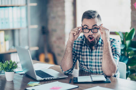 Portrait of handsome amazed surprised hipster wearing checkered shirt, he is touching his spectacles, sitting at the table is front of netbookの写真素材