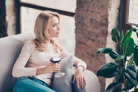 Charming, attractive, pretty, stylish woman, having glass with wine in hand sitting on couch with serious expression looking at window, thinking about somethingの写真素材