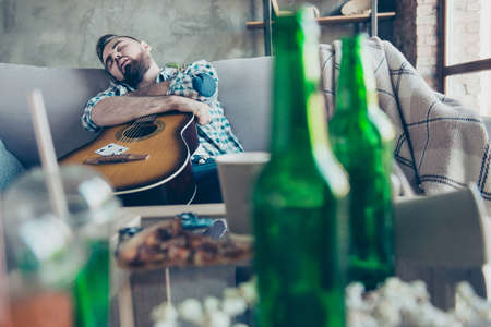 Drunk sleepy tired guy wearing checkered shirt is sleeping on a sofa and embracing an acoustic guitarの写真素材