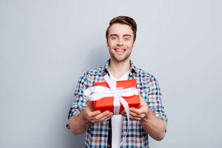 Portrait of cheerful, glad, smiling, positive gentelmen in checkered shirt giving, presenting, holding a gift in red package with white ribbon over grey backgroundの写真素材