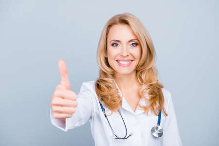 Happy, pretty, attractive, middle age medic worker in white coat showing thumb up to the camera over grey backgroundの写真素材