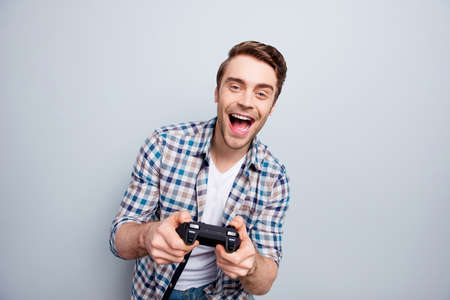 Portrait of crazy, cheerful, attractive, positive, very excited guy in checkered shirt holding joystick and playing video games with wide open mouth over grey backgroundの写真素材