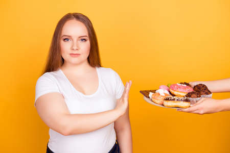 Will-powered woman wearing white tshirt is refusing to consume tasty delicious sweets on a plate, isolated on bright yellow backgroundの写真素材