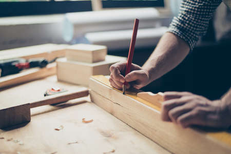 Cropped close up photo of foreman's hands holding a red pencil and measuring a wooden plank with a ruler on table, filing around on the tableの写真素材