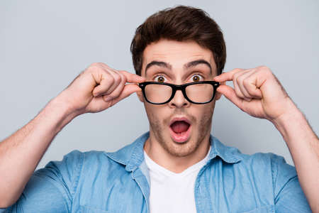 Close up portrait of astonished, shocked, attractive guy holding eyelets of glasses on his face with fingers, having wide open mouth, looking out spectaclesの写真素材