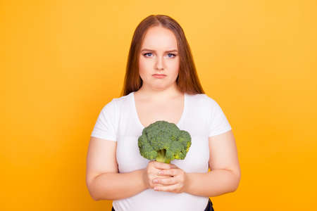 Portrait of unhappy upset sad frustrated woman dressed in white tshirt, she is holding green fresh raw broccoli, isolated on bright yellow backgroundの写真素材