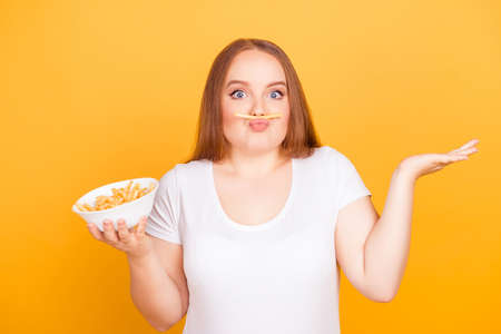 Happy wondered cheerful fatty lady is fooling around, she is making fake mustache using fried potatoes, isolated on bright yellow backgroundの写真素材