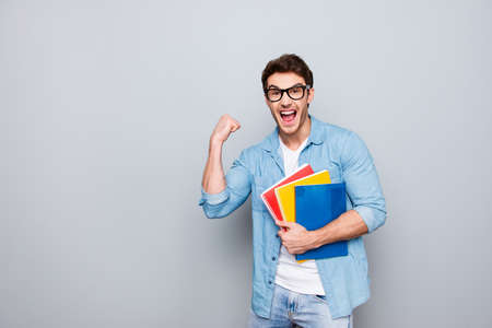 Portrait with copy space of cheerful, joyful, attractive guy with stubble having three colorful copybooks in arm, celebrating passed exams, graduation, isolated on grey backgroundの写真素材