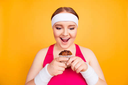 Funny excited cheerful fatty woman wearing sweat-band is holding a chocolate cupcake, looking with open mouth, she can put on weightの写真素材