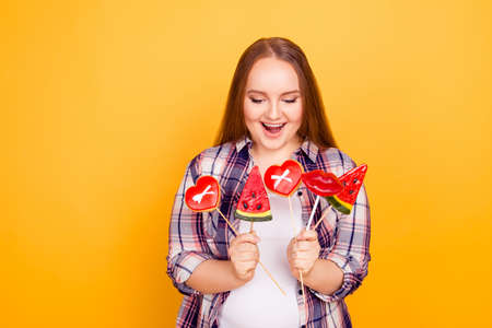 Portrait of excited with open mouth overweight woman clothed in checkered shirt, holding sweets and cookies on sticks isolated on yellow backgroundの写真素材